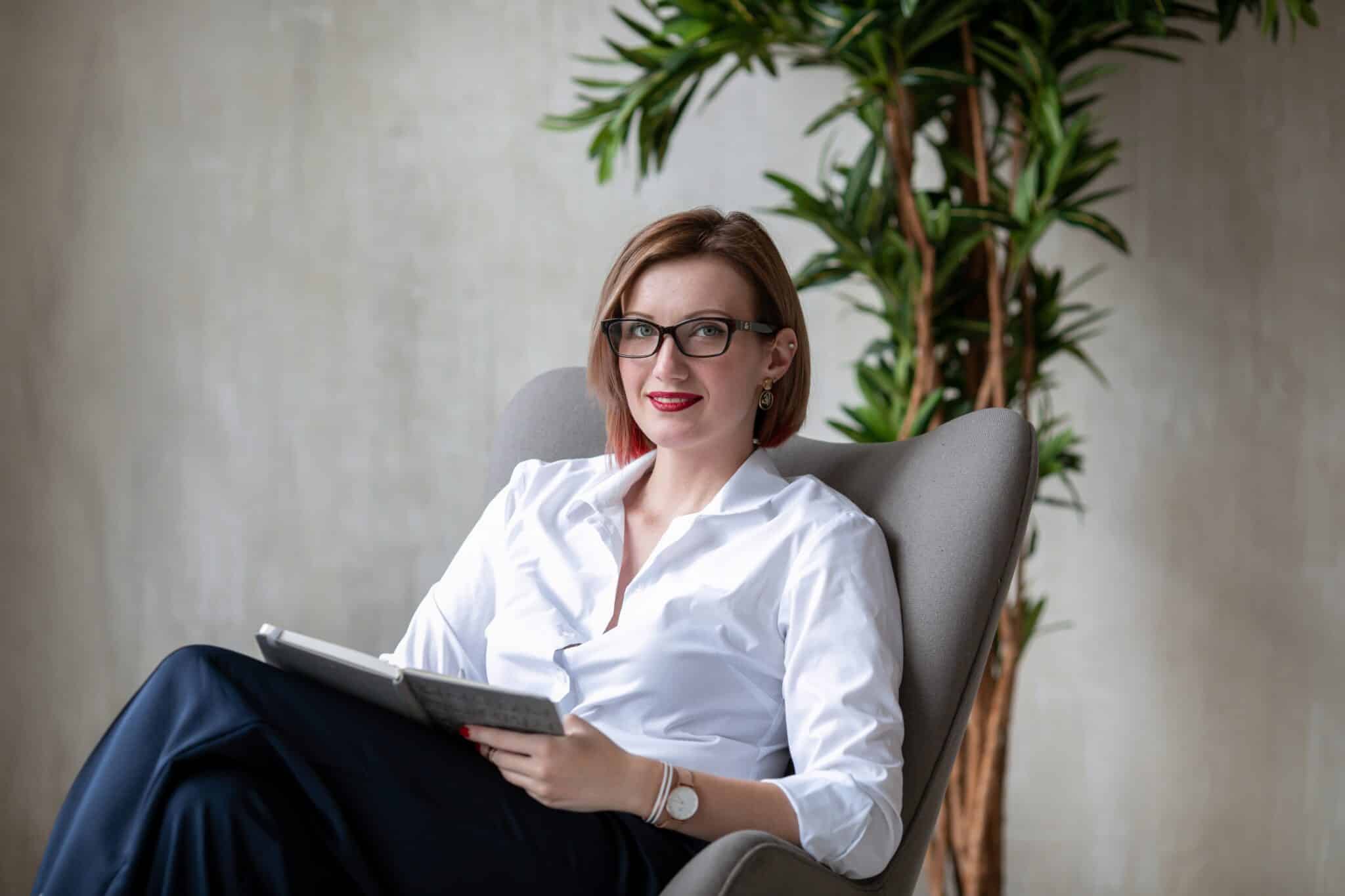 femme au cheveux court blond portant des lunettes assise sur un fauteuil avec un cahier en arrière plan une plante